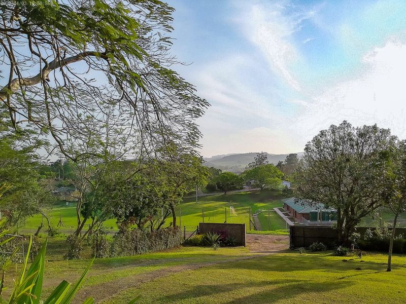 A scenic elevated view overlooking a green landscape with mature trees, fencing, and distant hills under a clear sky.