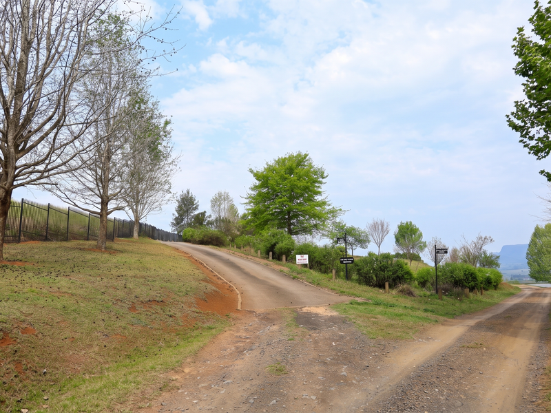 A view of the property entrance featuring a paved driveway, gravel access road, perimeter fencing, and established trees within a rural residential setting.