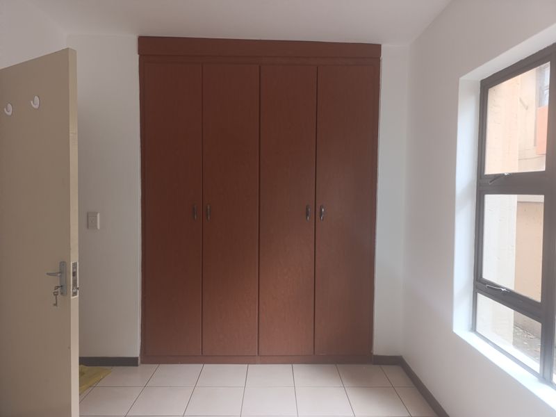 A clean, empty bedroom featuring built-in wooden wardrobes, neutral tiled flooring, and a large window providing natural light.