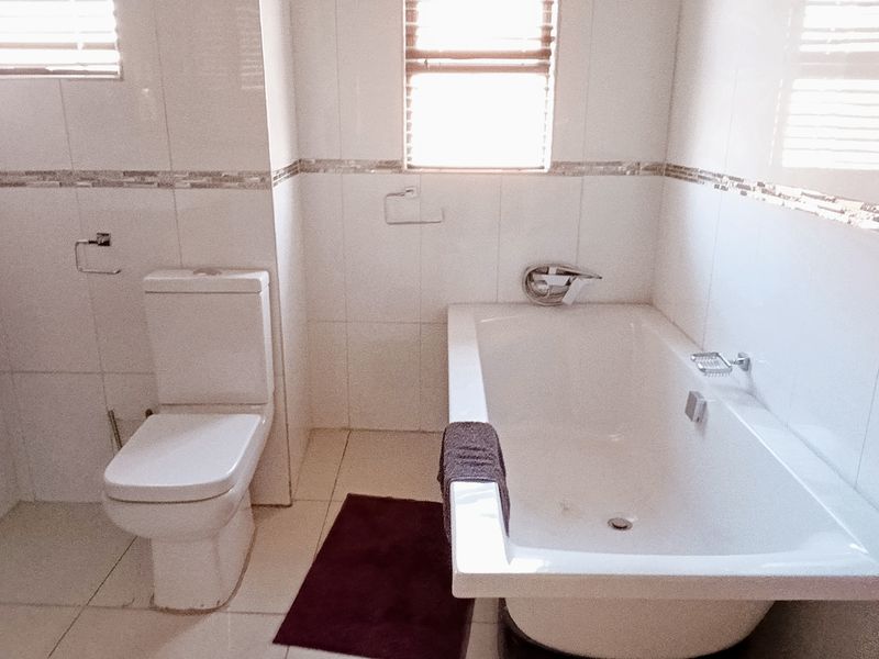 A clean, modern bathroom featuring white wall tiles, a built-in vanity, a standard toilet, and a bathtub with chrome fixtures.