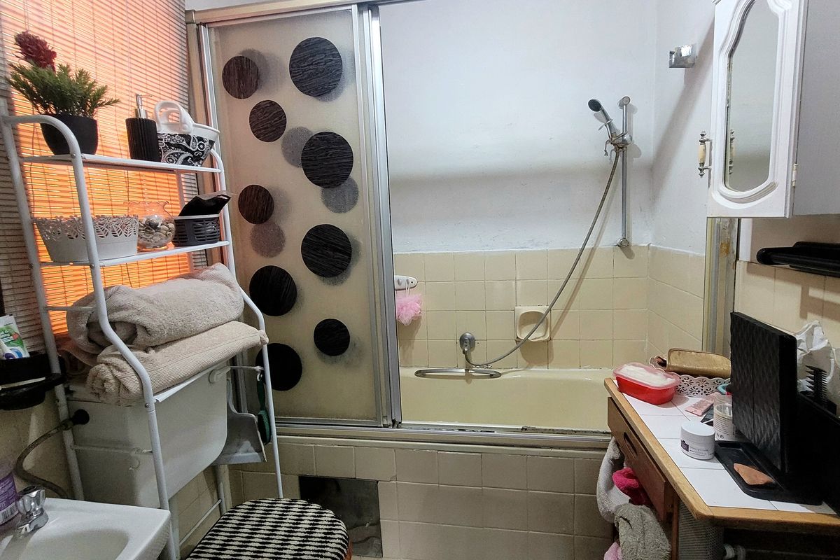 A dated bathroom featuring a bathtub with a sliding screen, vanity cabinet, and wall-mounted storage, requiring modernization of finishes and fixtures.