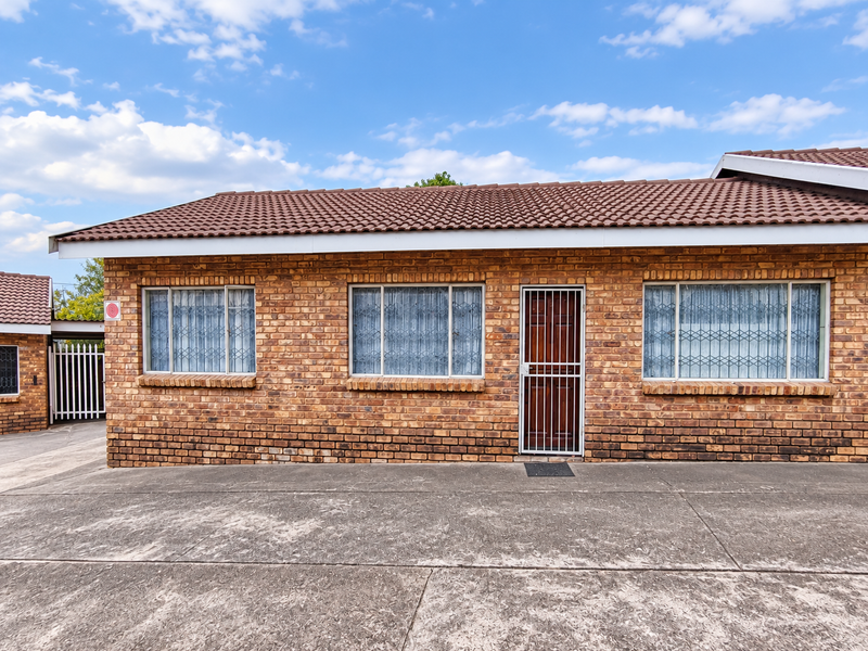 Front elevation of a face brick townhouse featuring a tiled roof, security gates, and a paved driveway area.