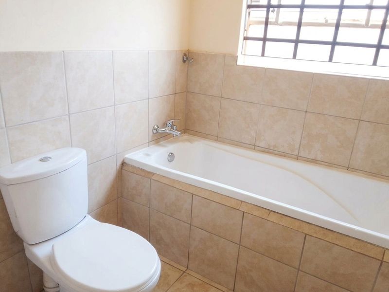 A functional bathroom featuring a built-in bathtub, ceramic tiling, and a standard toilet, with a window fitted with security bars.