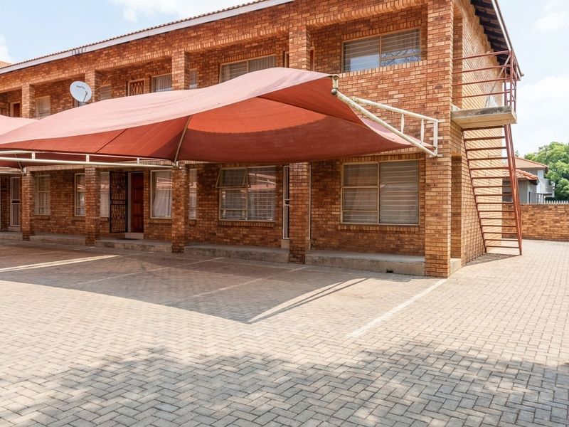 Exterior view of a face brick apartment complex featuring a paved parking area with a shade cloth carport and external metal staircase access.