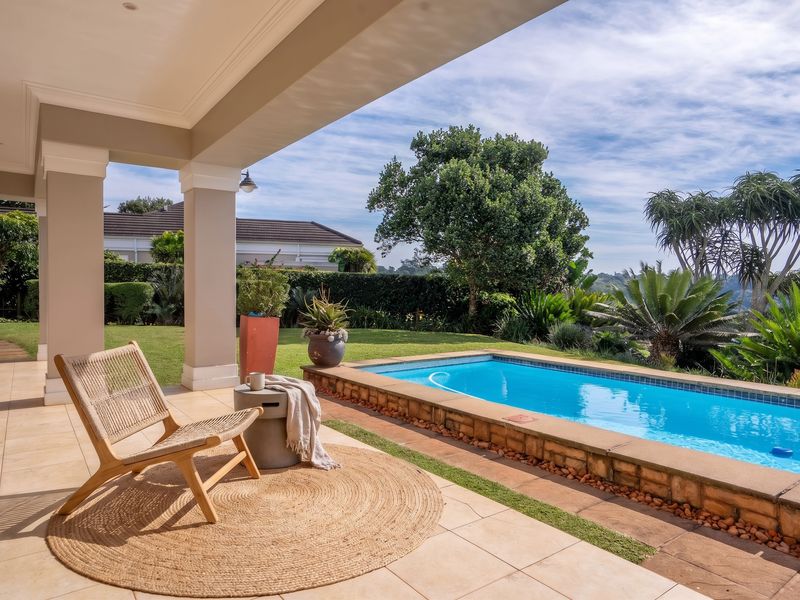 A spacious covered patio overlooking a raised swimming pool and a well-maintained garden, offering a serene outdoor living and entertainment space.