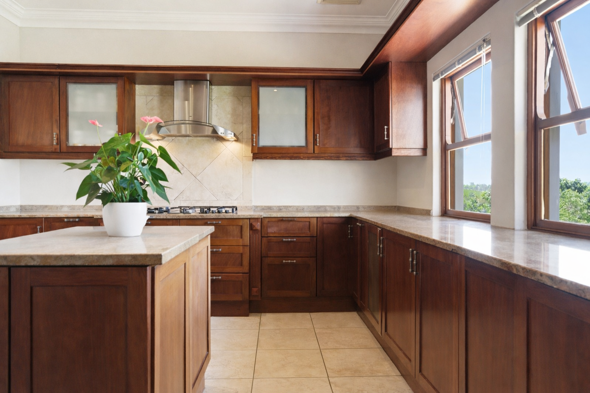 A well-maintained kitchen featuring rich wooden cabinetry, stone countertops, a central island, and a stainless steel extractor fan with natural light.