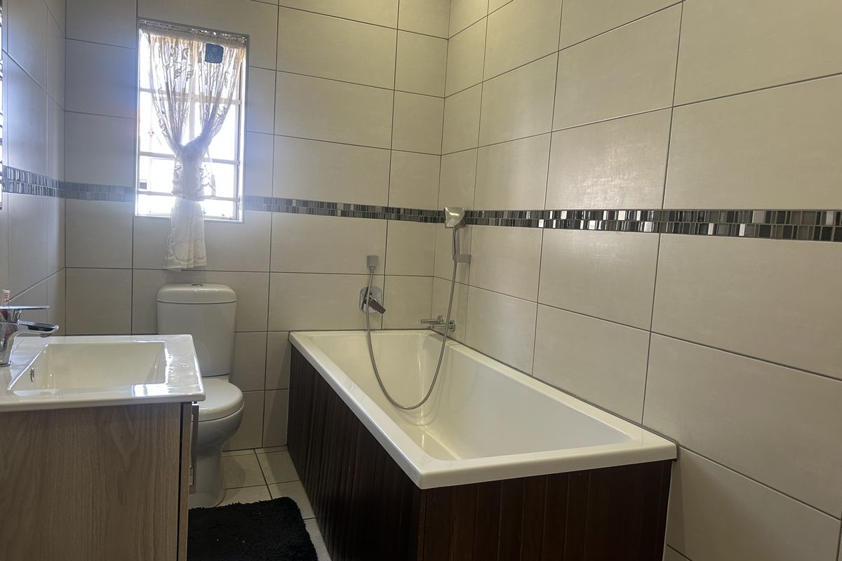 A clean, modern bathroom featuring a built-in bathtub with handheld shower mixer, a vanity cabinet, and neutral wall tiling with a decorative mosaic border.