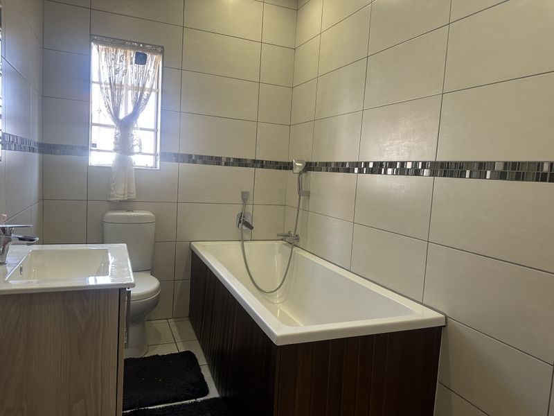 A clean, modern bathroom featuring a built-in bathtub with handheld shower mixer, a vanity cabinet, and neutral wall tiling with a decorative mosaic border.