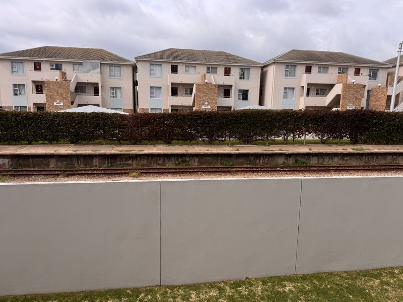 Exterior view of a multi-story apartment complex featuring brick facades, tiled roofing, and external staircases, situated behind a boundary wall and hedge.