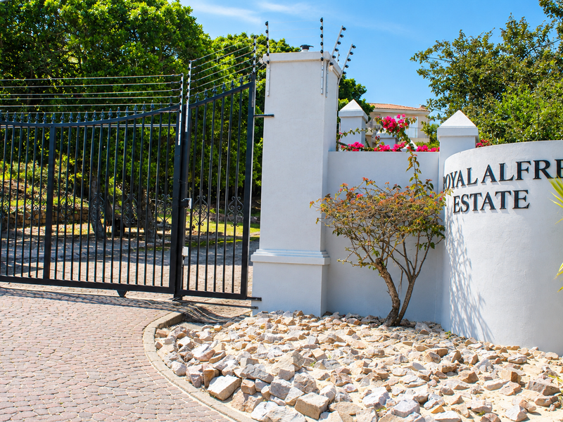 The entrance to Royal Alfred Estate featuring a secure wrought iron gate, electric fencing, and a well-maintained paved driveway.
