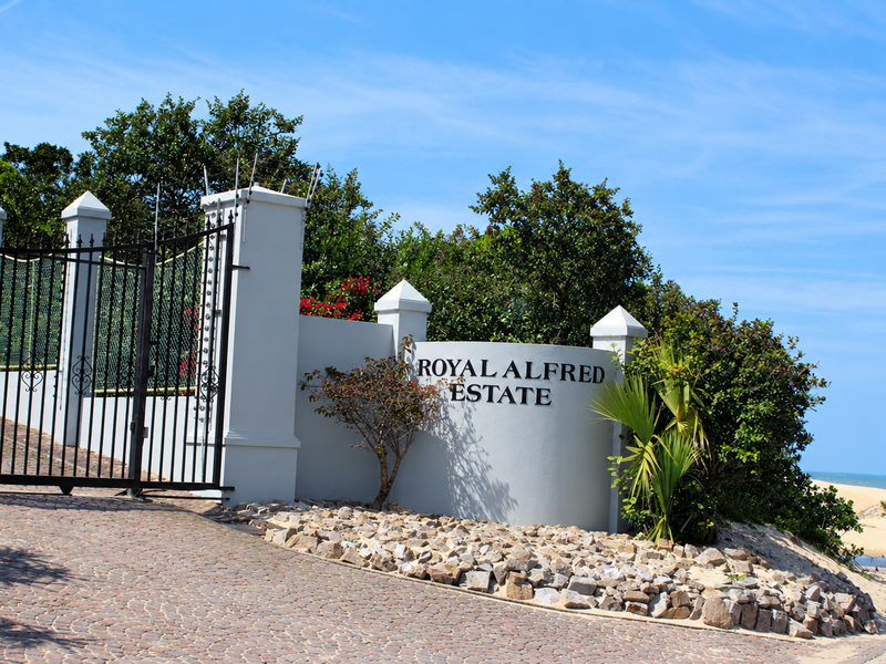 The entrance to Royal Alfred Estate featuring a secure gated access point, masonry pillars, and paved driveway with established landscaping.