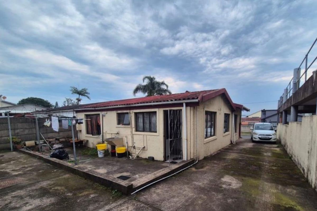 Rear view of the property featuring a concrete driveway, external wash basin, and perimeter walling, showing signs of weathering and maintenance requirements.