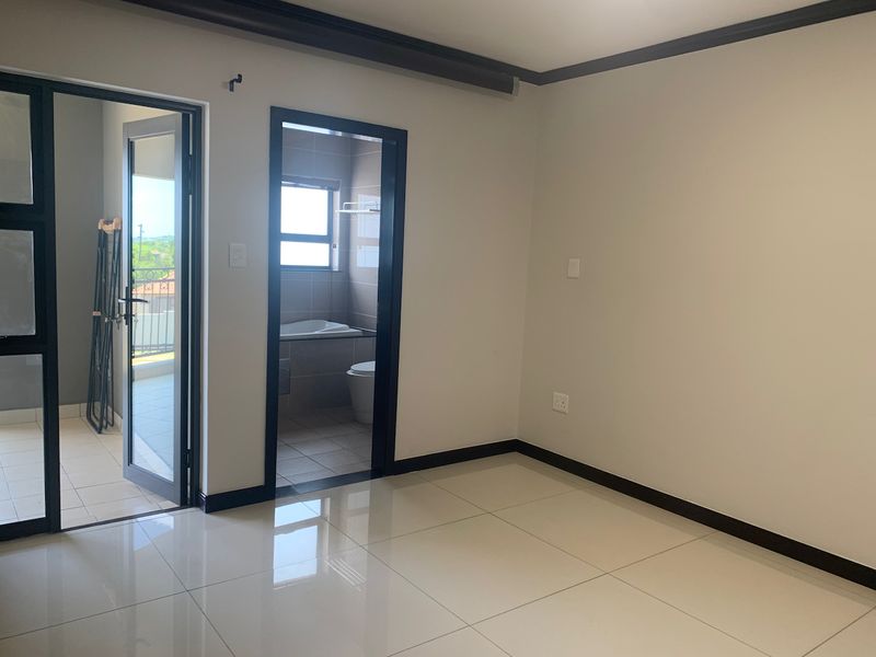 A bright, empty bedroom featuring polished tiled floors, dark contrasting door frames and skirting, with direct access to an en-suite bathroom and a private balcony.