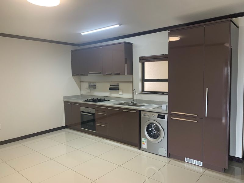 Modern kitchen featuring high-gloss brown cabinetry, integrated appliances, gas hob, built-in oven, and neutral tiled flooring in an open-plan layout.
