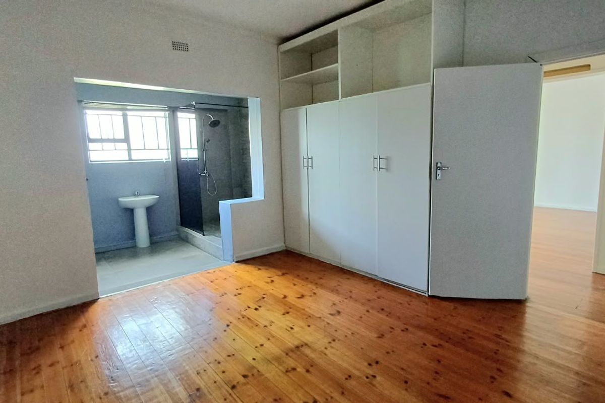 Spacious bedroom featuring polished hardwood flooring, ample built-in cupboard storage, and an open-plan en-suite bathroom with a shower and pedestal basin.