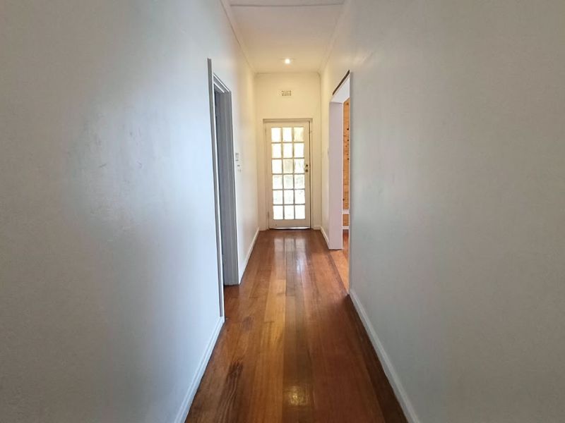 A bright, empty hallway featuring polished hardwood flooring and a classic white-framed French door at the end, providing access to the exterior.