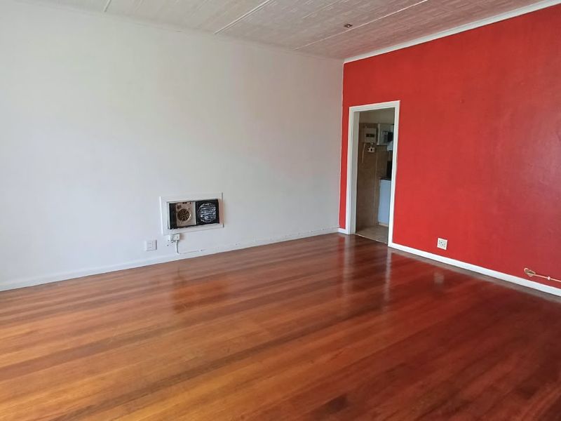An empty living room featuring polished hardwood flooring, a wall-mounted heater, and a distinctive red accent wall.