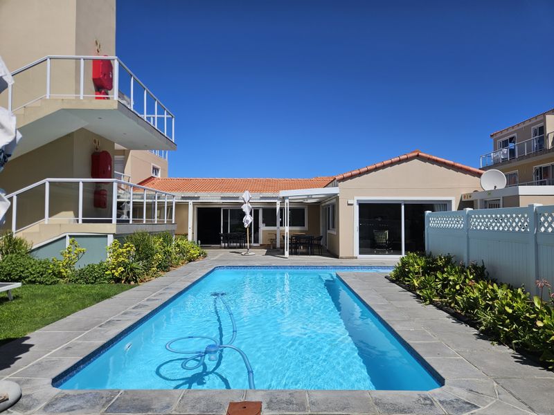 A well-maintained outdoor pool area featuring stone coping, surrounded by landscaped garden beds, white fencing, and access to the apartment building's ground floor.