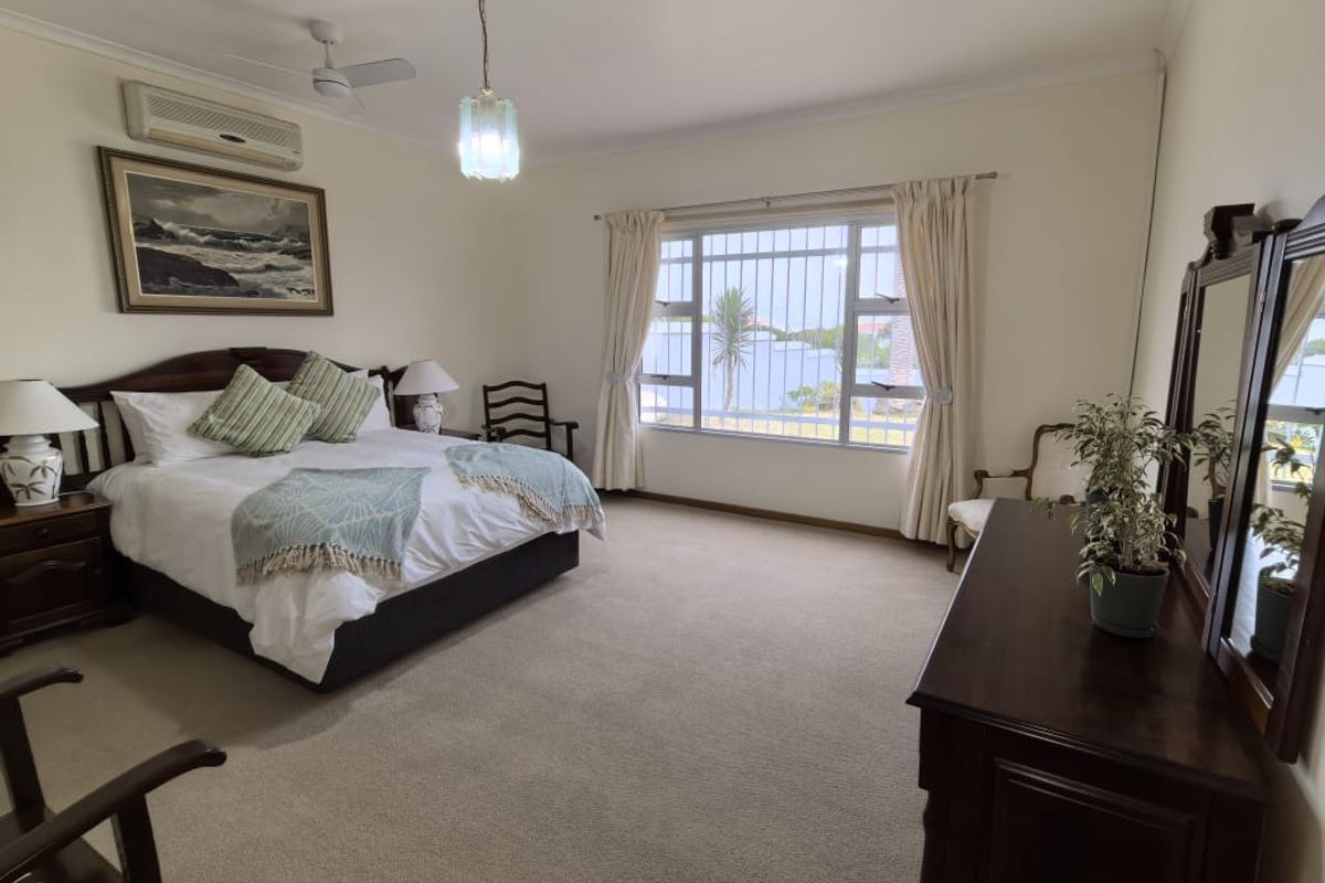 Spacious bedroom featuring neutral carpeting, a ceiling fan, and a wall-mounted air conditioning unit, with large windows fitted with security bars.