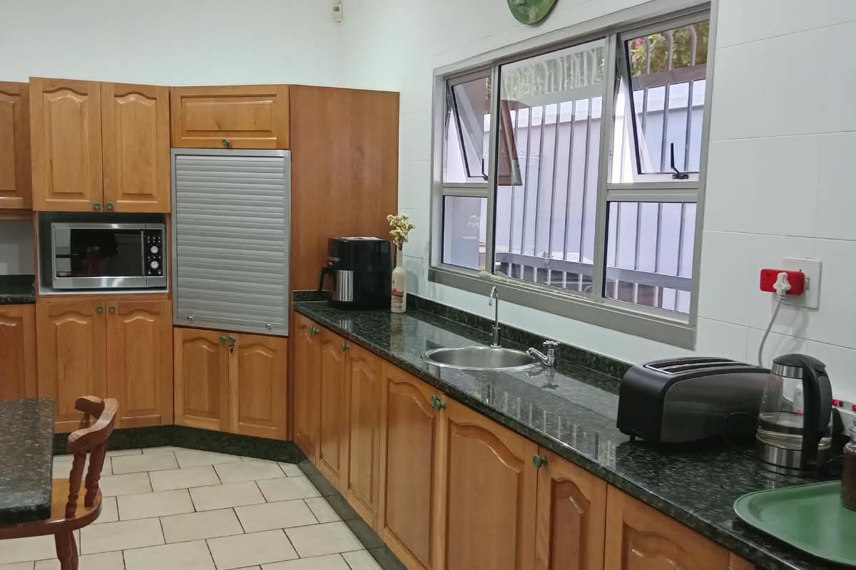 A functional kitchen featuring wooden cabinetry, granite countertops, a stainless steel sink, and tiled flooring, offering a practical workspace with natural light.