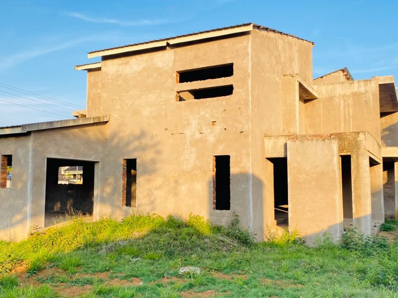 A partially constructed residential house showing the exterior shell with plastered walls and open window and door frames on a vacant plot.