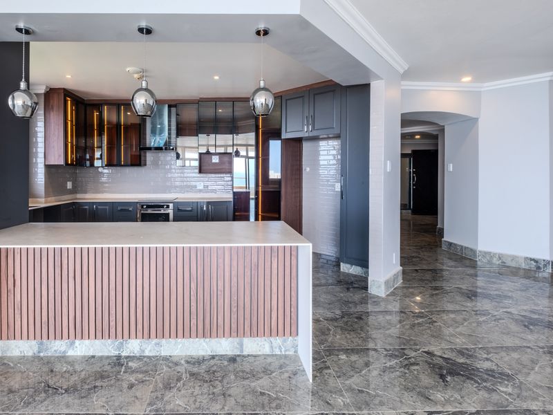 Modern kitchen featuring dark cabinetry, a central island with wood-slat detailing, built-in appliances, and polished marble flooring throughout the open-plan space.
