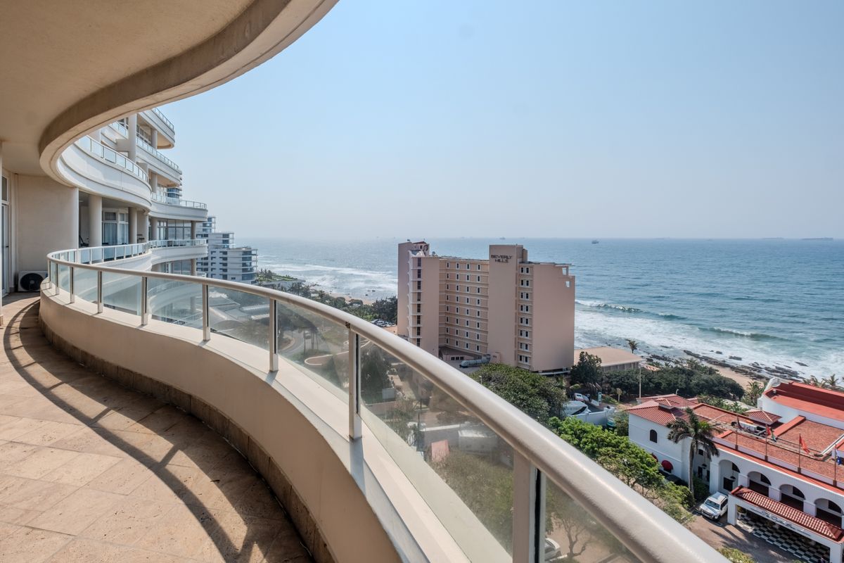 A spacious, curved balcony featuring a glass balustrade and tiled flooring, offering expansive ocean and coastal views.