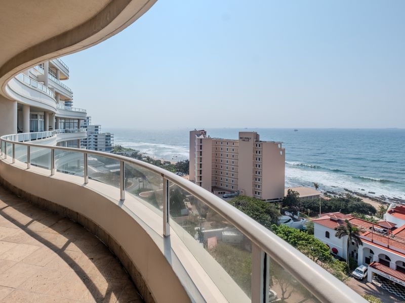 A spacious, curved balcony featuring a glass balustrade and tiled flooring, offering expansive ocean and coastal views.