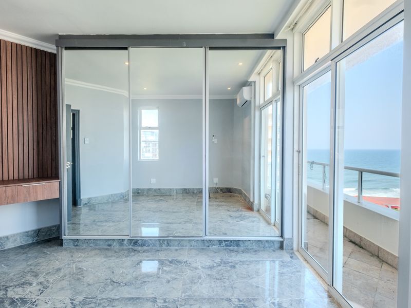 Spacious bedroom featuring large mirrored sliding wardrobes, marble-look tiled flooring, wall-mounted air conditioning, and direct access to a balcony with ocean views.