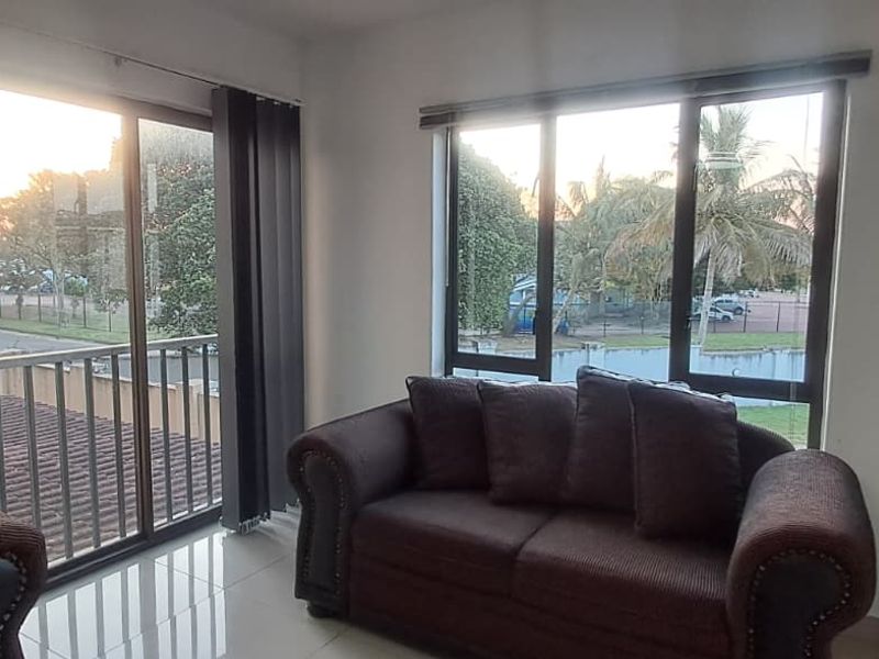 A bright living room featuring large windows and a sliding glass door leading to a balcony, with neutral tiled flooring throughout.