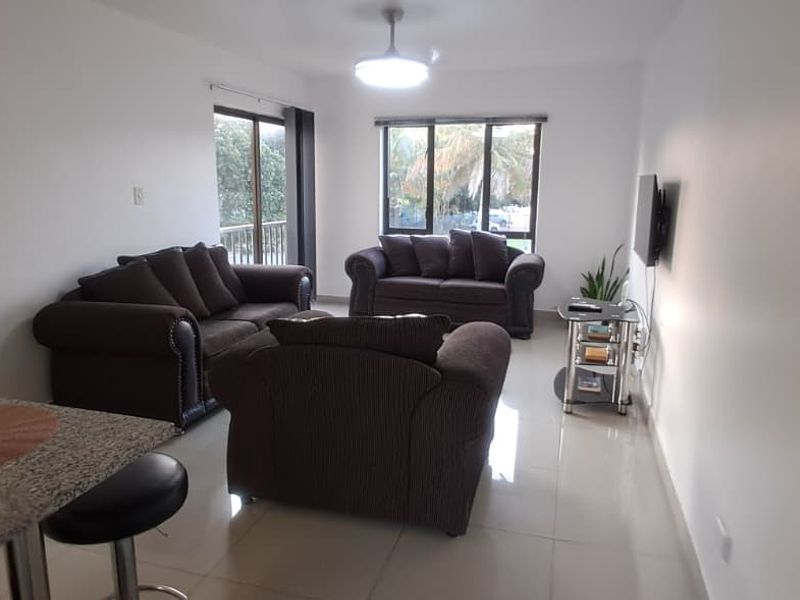 A bright and spacious living room featuring neutral tiled flooring, a ceiling fan, and large windows providing natural light and access to a balcony.
