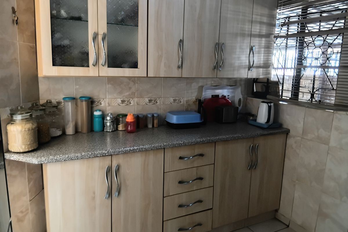 A functional kitchen featuring light wood-tone cabinetry, granite-look laminate countertops, and neutral ceramic wall and floor tiling with security bars on the window.