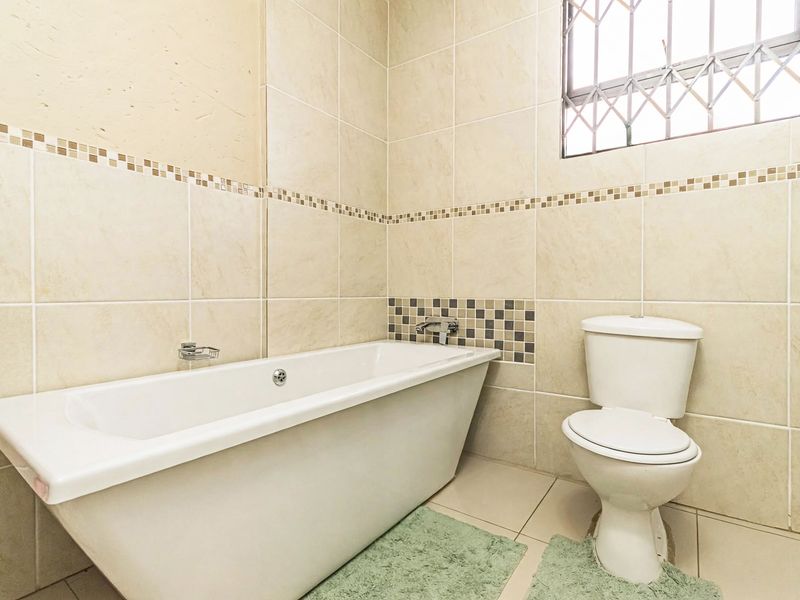 A functional bathroom featuring a bathtub, toilet, and neutral wall tiling with decorative mosaic accents and a security-fitted window.