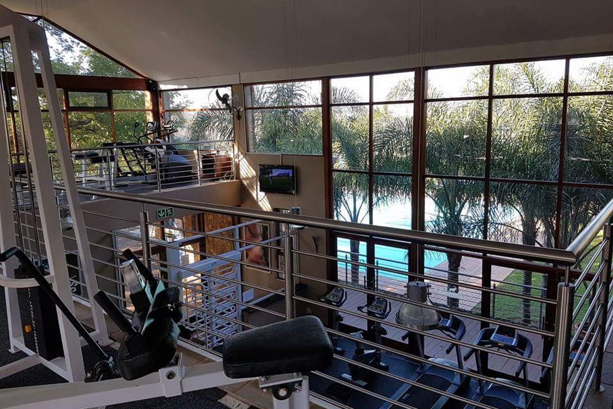 A spacious, light-filled mezzanine gym featuring high vaulted ceilings, extensive floor-to-ceiling windows, and a modern stainless steel balustrade overlooking the pool area.