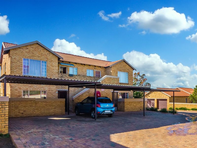 A well-maintained face brick apartment complex featuring a paved driveway, covered carports, and a secure boundary wall.