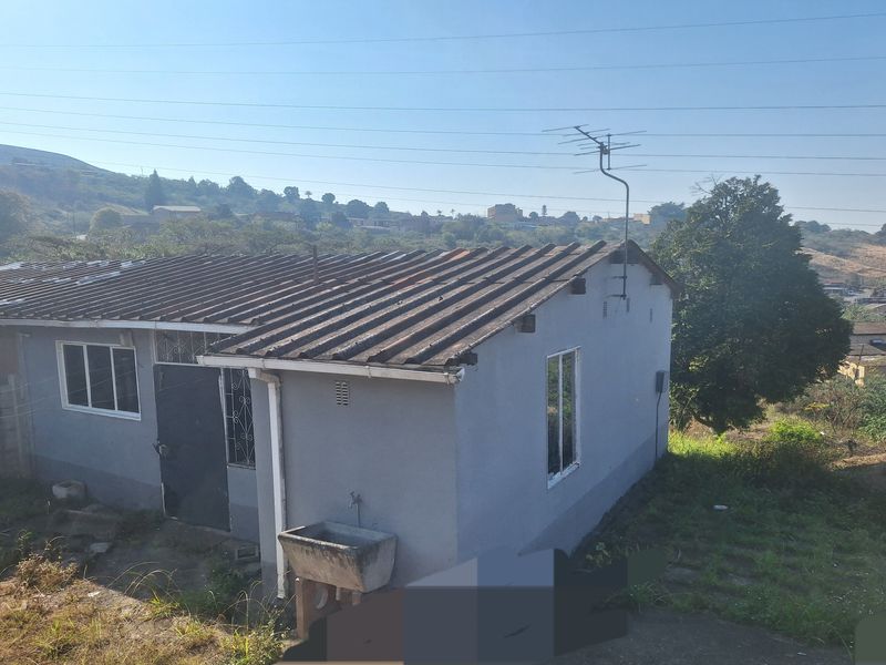 Rear view of a single-story house featuring a corrugated roof, external wash basin, and security gate, set against a hillside landscape.