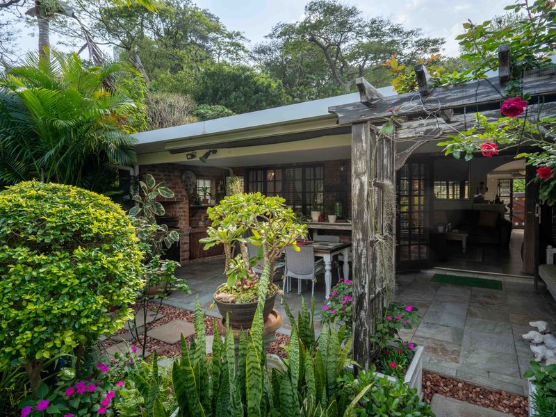 A covered patio area featuring stone paving, a built-in brick braai, and a wooden pergola, offering a seamless transition to the garden.