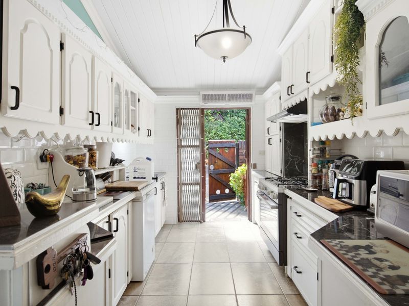 A galley-style kitchen featuring white cabinetry, dark countertops, subway tile backsplash, and direct access to the rear exterior through a security gate.