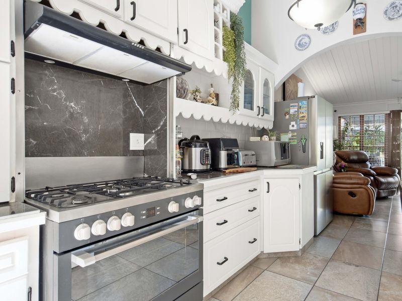 A functional kitchen featuring a gas stove, oven, extractor fan, stone splashback, and ample built-in cabinetry, flowing into an adjacent living area.