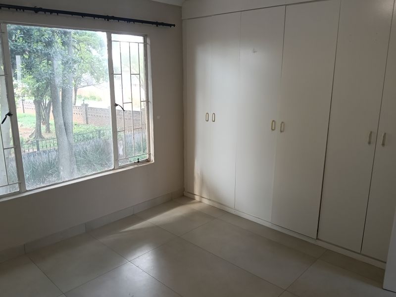 An empty bedroom featuring tiled flooring, built-in cupboards, and a large window with security bars overlooking the garden.