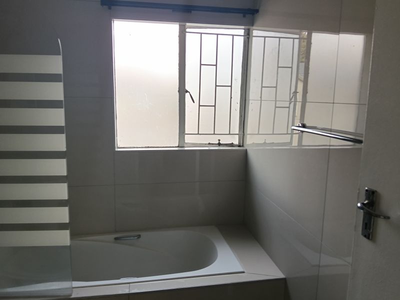 A functional bathroom featuring a built-in bathtub with a glass screen, tiled walls, and a window with security bars.