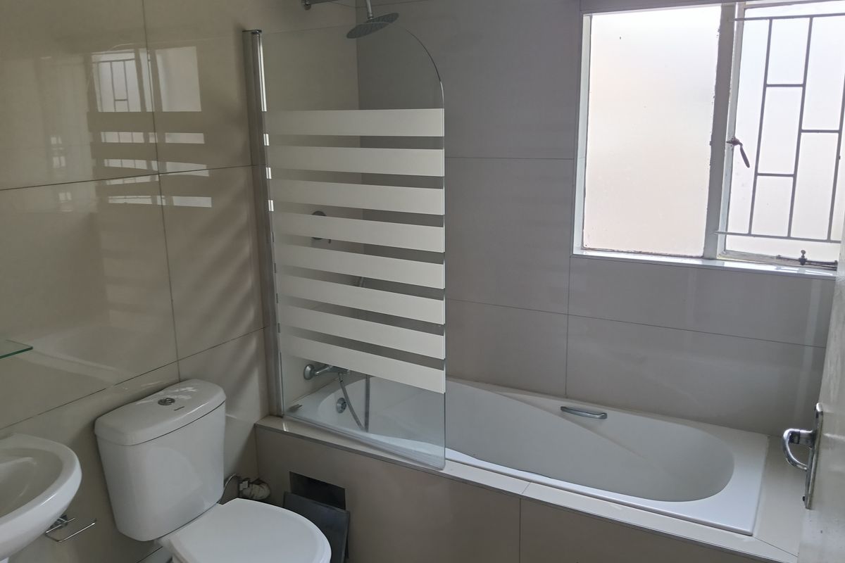 A clean, functional bathroom featuring a bathtub with a glass shower screen, pedestal basin, and tiled walls with security bars on the window.