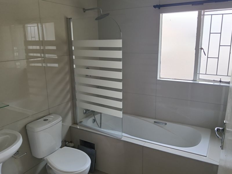 A clean, functional bathroom featuring a bathtub with a glass shower screen, pedestal basin, and tiled walls with security bars on the window.