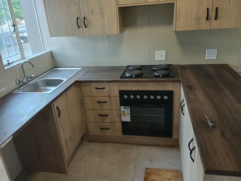 A modern, compact kitchen featuring light wood-finish cabinetry, dark laminate countertops, a built-in oven, electric hob, and a stainless steel sink.