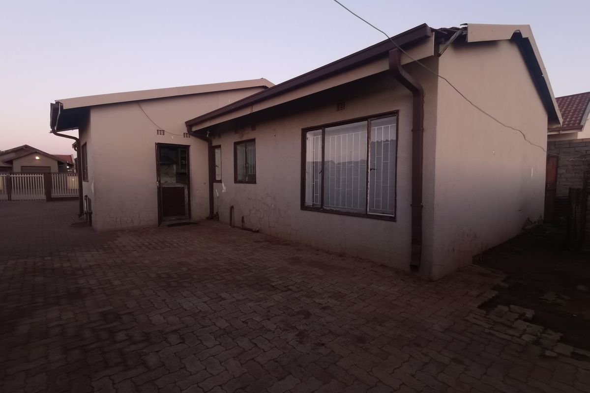 Rear exterior view of the house featuring paved ground, security bars on windows, and visible wear to the exterior wall plaster and roofline.