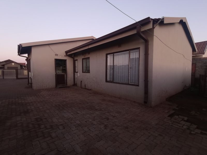Rear exterior view of the house featuring paved ground, security bars on windows, and visible wear to the exterior wall plaster and roofline.