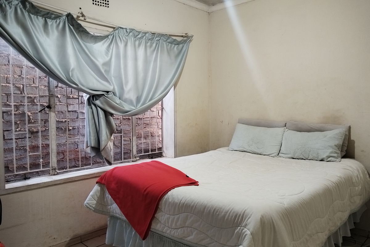 A bedroom featuring tiled flooring, security bars on the window, and a ventilation brick, currently showing signs of wear on the walls and ceiling.