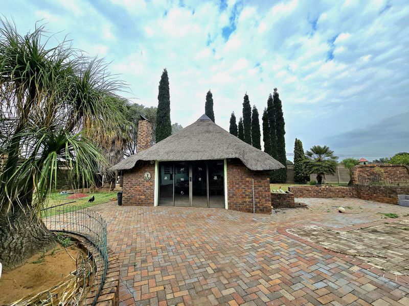A rear exterior view featuring a thatched roof structure with brick chimney, sliding glass doors, and extensive brick paved patio area.