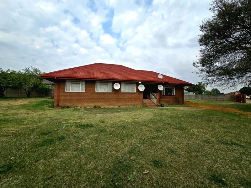 A detached single-story brick house featuring a prominent red tiled roof, metal-framed windows, and a large surrounding lawn area.