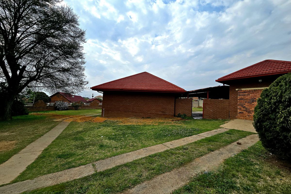 Rear view of the property featuring a brick exterior, red tiled roof, and a spacious garden area with concrete pathways and driveway.